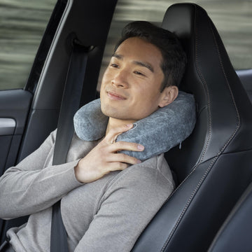Ein Mann sitzt entspannt in einem Auto, trägt einen Sicherheitsgurt und benutzt ein Reise-Nacken-Massagekissen mit Vibrationsmassage in Grau.