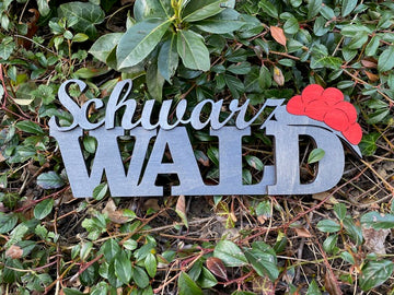 Ein Schwarzwald-Schriftzug mit rotem Bollenhuthut-Motiv ruht auf grünen Blättern und bringt einzigartigen Schwarzwald-Charme mit.