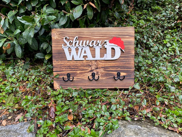 Das Schwarzwald-Schlüsselbrett „Christine“ ist ein im Schwarzwald-Stil gefertigtes Schlüsselbrett aus Kiefernholz mit vier Haken, das auf grünem Blätterboden in der Nähe von Büschen abgebildet ist.