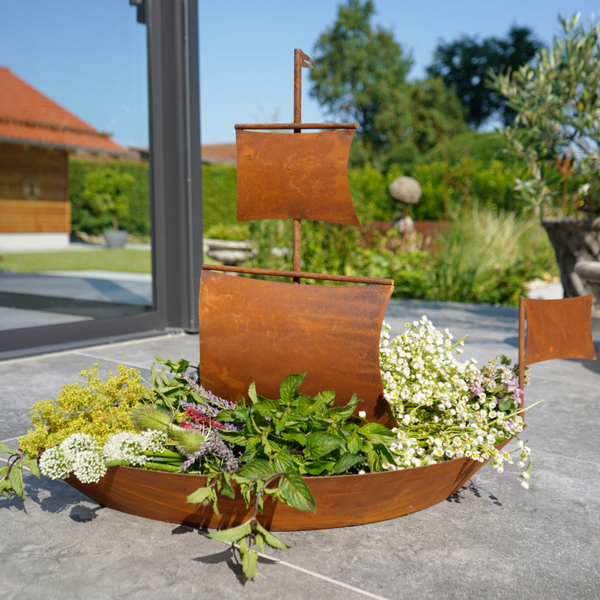 Das Edelrost-Schiff zum Bepflanzen, ein metallener, bootförmiger Pflanzkübel in Vintage-Rostoptik, wird mit Kräutern und Blumen gefüllt und auf einer Terrasse in der Nähe von Glastüren aufgestellt, umgeben von üppigem Grün.