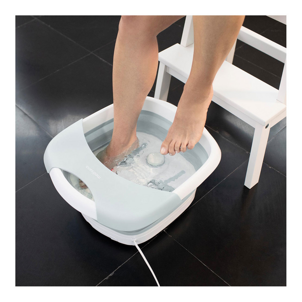 Eine Person genießt eine entspannende Schüttelmassage, während sie ihre Füße in einem faltbaren Fußsprudelbad badet und auf einem weißen Stuhl auf einem schwarzen Fliesenboden sitzt.