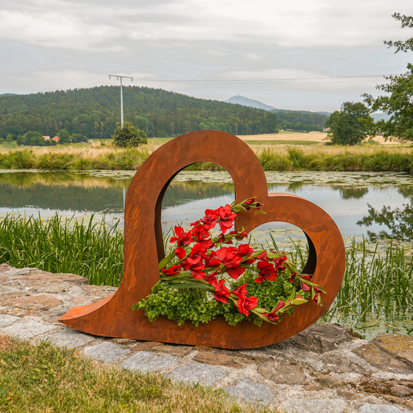 Das Edelrost 2er Set Herz zum Bepflanzen sind rustikale, herzförmige Pflanzgefäße im Vintage-Rost-Look, die mit roten Blumen geschmückt und neben einem Teich mit grünen Feldern im Hintergrund präsentiert werden.