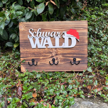 Das Schwarzwald-Schlüsselbrett „Christine“ ist ein im Schwarzwald-Stil gefertigtes Schlüsselbrett aus Kiefernholz mit vier Haken, das auf grünem Blätterboden in der Nähe von Büschen abgebildet ist.