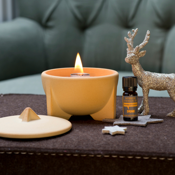 Ein brennender SCHMELZFEUER® INDOOR CERANATUR®-Kerzenhalter, eine Flasche mit ätherischem Öl, Sternornamente und ein glitzerndes Rentier sorgen auf einer braunen Oberfläche für eine gemütliche Aromatherapie-Atmosphäre.