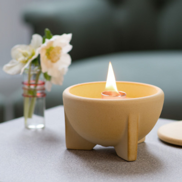 Die gelbe Keramikkerze SCHMELZFEUER® INDOOR CERANATUR® leuchtet auf einem Tisch, ideal für die Aromatherapie, mit weißen Blumen in einer kleinen Vase im Hintergrund.