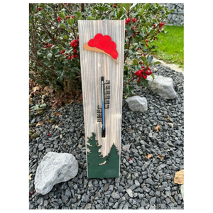 Das Schwarzwald-Thermometer aus weißem Kiefernholz begeistert mit Kiefernbaum- und roter Wolkendekoration, präsentiert auf grauem Kies und Pflanzen im Hintergrund.