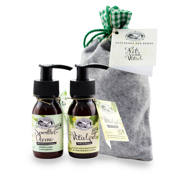 Zwei kleine Fläschchen Fit & Vital-Set Kräutercreme mit Pumpverschluss stehen neben einem grauen Stoff-Geschenkbeutel mit grün-kariertem Band. Die wohltuende Formel mit Kampfer und Menthol sorgt für ein erfrischendes Erlebnis.