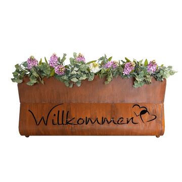 Das Edelrost Pflanzengefäß Bregenz - Willkommen hat einen braunen Rost-Look mit rosa Blumen und „Willkommen“ auf der Vorderseite, perfekt, um Ihrem Raum den Charme des Bayerischen Waldes zu verleihen.