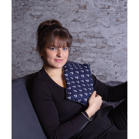 Eine Frau sitzt auf einem Sofa, lächelt in die Kamera und hält ein marineblaues Kirschkernkissen, 3 Kammern mit maritimem Design, an ihre Brust.