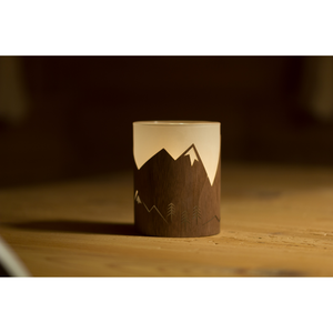 Ein beleuchtetes Windlicht Winterlicht mit Berg- und Baummotiv ruht in seiner Halterung auf einer Oberfläche aus Nussbaumholz.