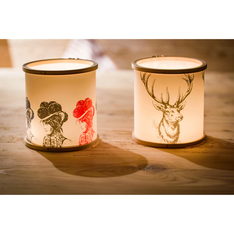 Zwei Windlicht Stitch Light-Laternen mit Illustrationen im Vintage-Stil von Menschen mit Hüten und einem Hirsch leuchten wunderschön auf einer Holzoberfläche.