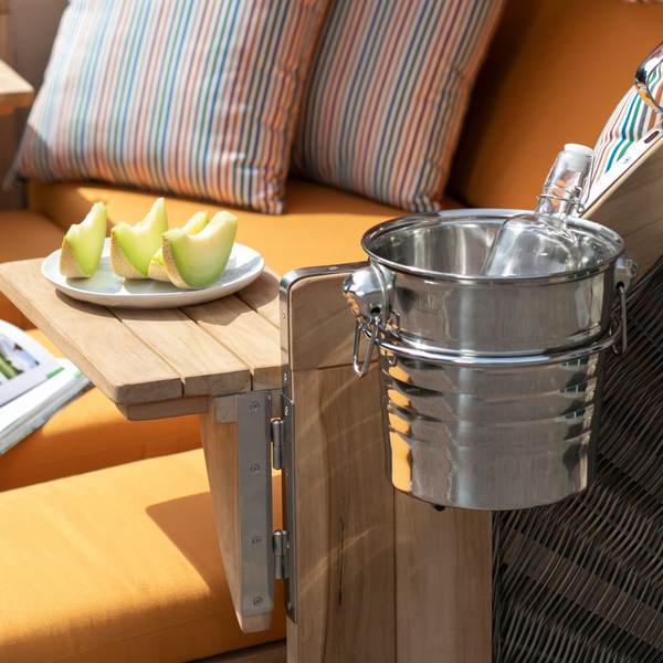 Ein gemütlicher Sitzplatz im Freien mit orangefarbenen Kissen, gestreiften Kissen, Melonenscheiben und dem Strandkorb-Getränkekühler, der Ihre Flasche kalt hält – ein witterungsbeständiges Luxus-Accessoire zum stilvollen Entspannen.