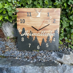 Die Schwarzwalduhr ist eine quadratische Kiefernholzuhr mit der Aufschrift „Heimatliebe“, Bäumen und einem Hirsch, die im Freien zwischen Felsen und grünem Efeu ausgestellt ist.