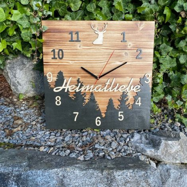 Die Schwarzwalduhr ist eine quadratische Kiefernholzuhr mit der Aufschrift „Heimatliebe“, Bäumen und einem Hirsch, die im Freien zwischen Felsen und grünem Efeu ausgestellt ist.