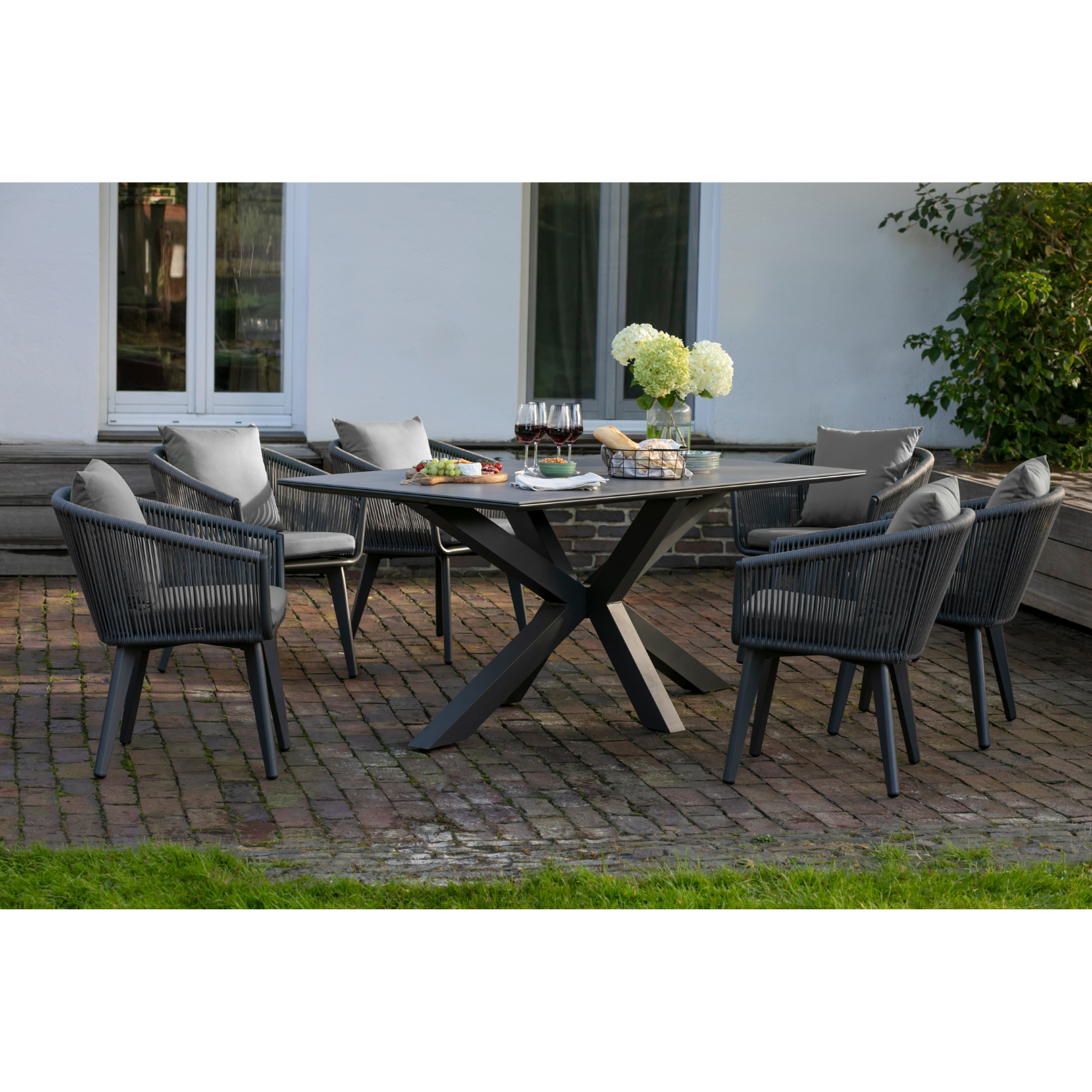 Modernes Gartenmöbelset Rope & Alu mit Tisch und sechs Stühlen, dekorativ mit Blumen, Wein und Speisen auf einer stilvollen Backsteinterrasse arrangiert.
