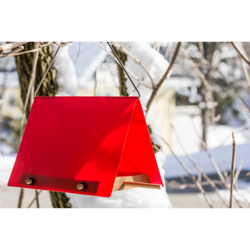 Ein rotes Vogelfutterhaus mit Edelstahlbügel hängt an einem verschneiten Ast unter einem strahlenden Winterhimmel.