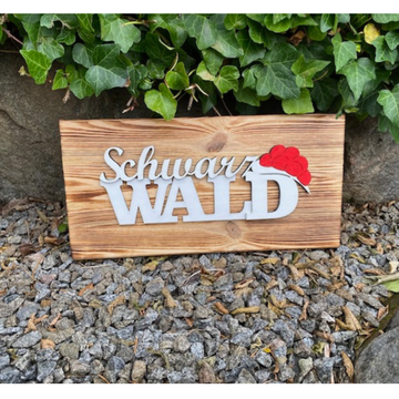 Das Schwarzwald Dekobrett „Franzi“ und „Heidi“ aus Kiefernholz mit Black Forest Schriftzug und rotem Hut steht auf Kies vor grünem Efeu.