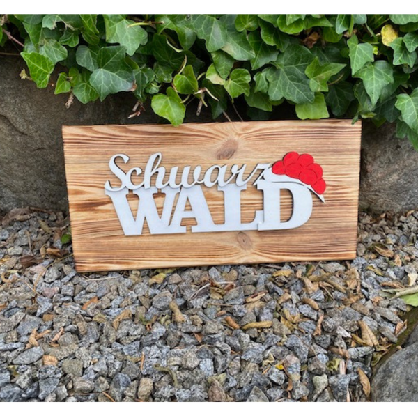 Das Schwarzwald Dekobrett „Franzi“ und „Heidi“ aus Kiefernholz mit Black Forest Schriftzug und rotem Hut steht auf Kies vor grünem Efeu.
