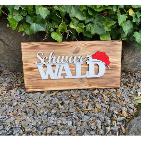 Das Schwarzwald Dekobrett „Franzi“ und „Heidi“ aus Kiefernholz mit Black Forest Schriftzug und rotem Hut steht auf Kies vor grünem Efeu.