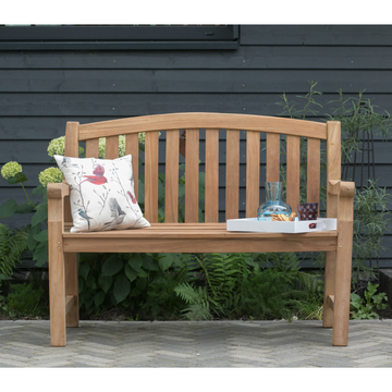 Die Gartenbank Swindon aus Naturholz präsentiert sich mit ergonomischer Form; Darauf liegt ein Vogel-Kissen, daneben ein Tablett mit Glaskaraffe und Tassen – stimmungsvoll arrangiert vor einer dunklen Außenwand.