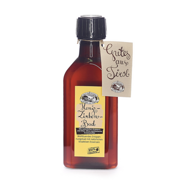 Braune Flasche mit gelbem Etikett und „Grüße aus Tirol“-Anhänger enthält Honig Zirben Bad Badeessenz mit Imkerhonig für luxuriöse Hautpflege und verbesserte Elastizität.