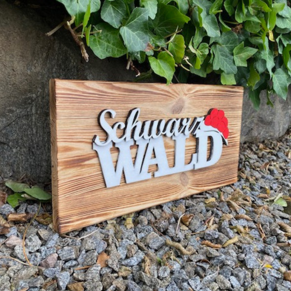 Das Schwarzwald Dekobrett „Franzi“ und „Heidi“ zeigt einen Schwarzwälder Schriftzug und einen roten Hut, dargestellt auf Kies neben Efeu und Felsen.
