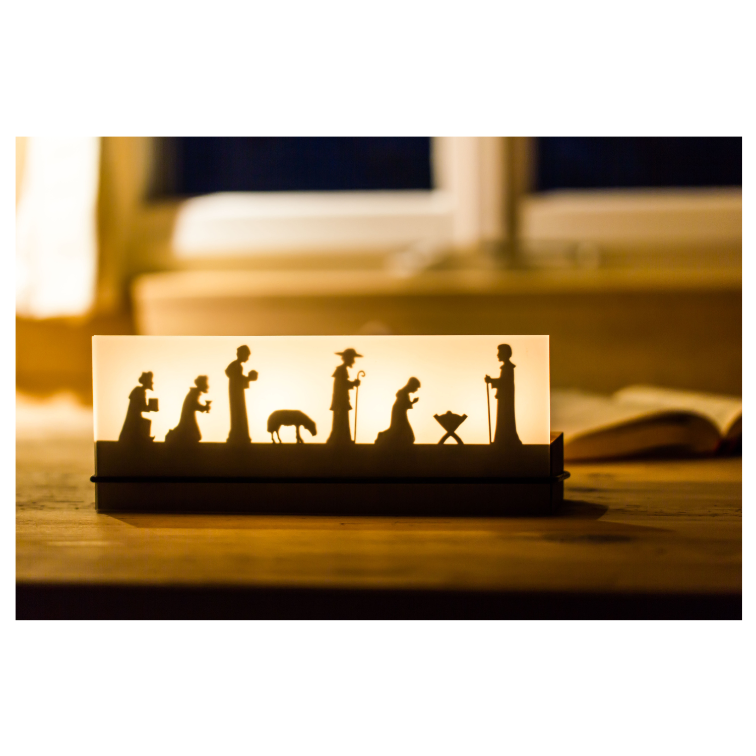 Der Teelichtleuchter Schattenspiel 4er Krippe beleuchtet eine Krippensilhouette auf einem Holztisch am Fenster und stellt so die Weihnachtsgeschichte wunderschön dar.