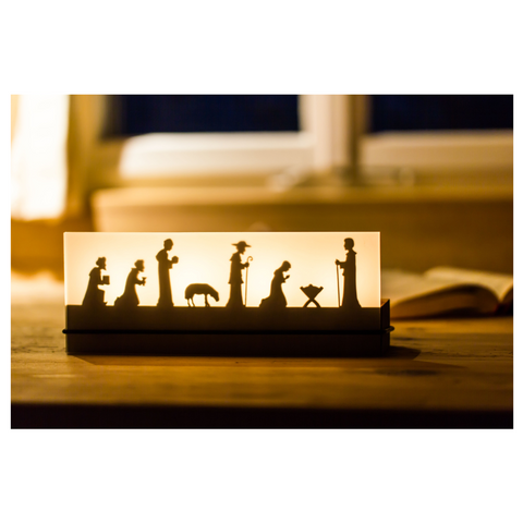Der Teelichtleuchter Schattenspiel 4er Krippe beleuchtet eine Krippensilhouette auf einem Holztisch am Fenster und stellt so die Weihnachtsgeschichte wunderschön dar.