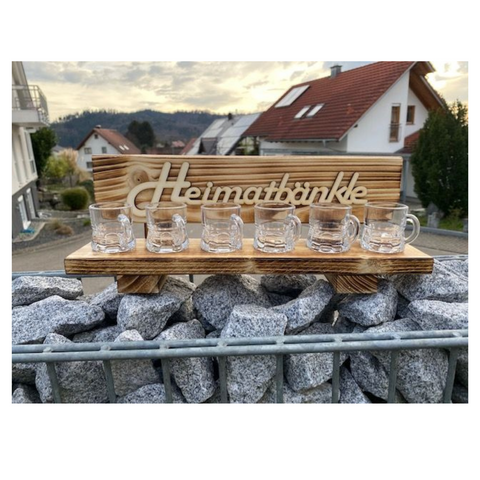 Sechs Schnapsgläser sind auf dem Schwarzwald 6er Schnapsbänkle „Edelbert“ arrangiert, im Freien mit charmanten Häusern im Hintergrund.
