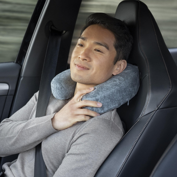 Ein Mann sitzt entspannt in einem Auto, trägt einen Sicherheitsgurt und benutzt ein Reise-Nacken-Massagekissen mit Vibrationsmassage in Grau.