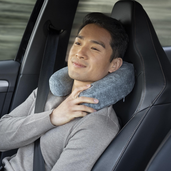 Ein Mann sitzt entspannt in einem Auto, trägt einen Sicherheitsgurt und benutzt ein Reise-Nacken-Massagekissen mit Vibrationsmassage in Grau.