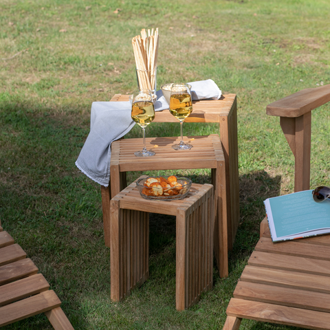 Zwei Holzstühle, Getränke, Snacks, ein Buch und eine Sonnenbrille liegen auf dem Beistelltischen 3er-Set, das im Freien auf grünem Gras steht.