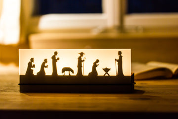 Der Teelichtleuchter Schattenspiel 4er Krippe beleuchtet eine Krippensilhouette auf einem Holztisch am Fenster und stellt so die Weihnachtsgeschichte wunderschön dar.
